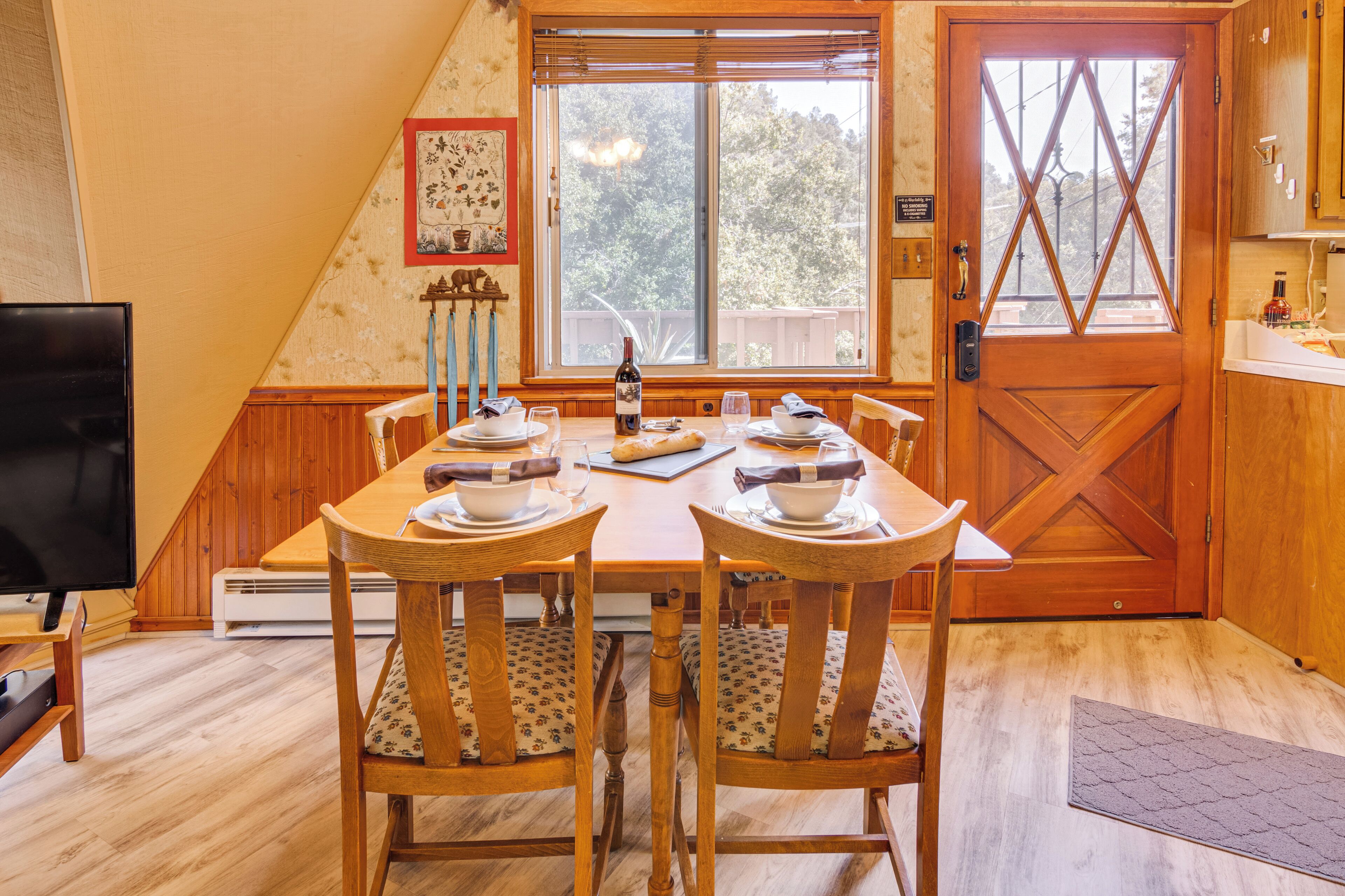 A dining area with a wooden table set for four, a television, and a window with a view of trees.