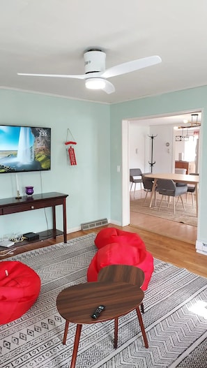 Een woonkamer met een flatscreen televisie, een plafondventilator, een eethoek met stoelen en een kamerbreed tapijt met een patroon.