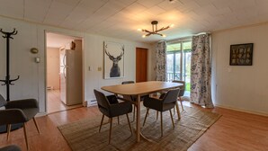 A dining area with a wooden table, four chairs, a chandelier, and a view of the outdoors through a window with floral curtains.