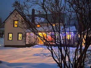 Een huis met een sneeuwbedekte grond, een zichtbare schoorsteen en een verlicht raam.