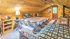 Ein gemütliches Schlafzimmer im Dachgeschoss mit Holzbalkendecke, einem Bett mit gemustertem Überwurf, einem kleinen Schreibtisch mit Lampe und einem Fenster mit Blick ins Freie.