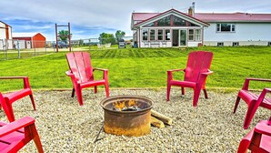 A fire pit with red chairs arranged around it, set on a gravel surface with a grassy field and a house in the background. - Stunning Wisconsin Retreat on Active Farm! (Arena)