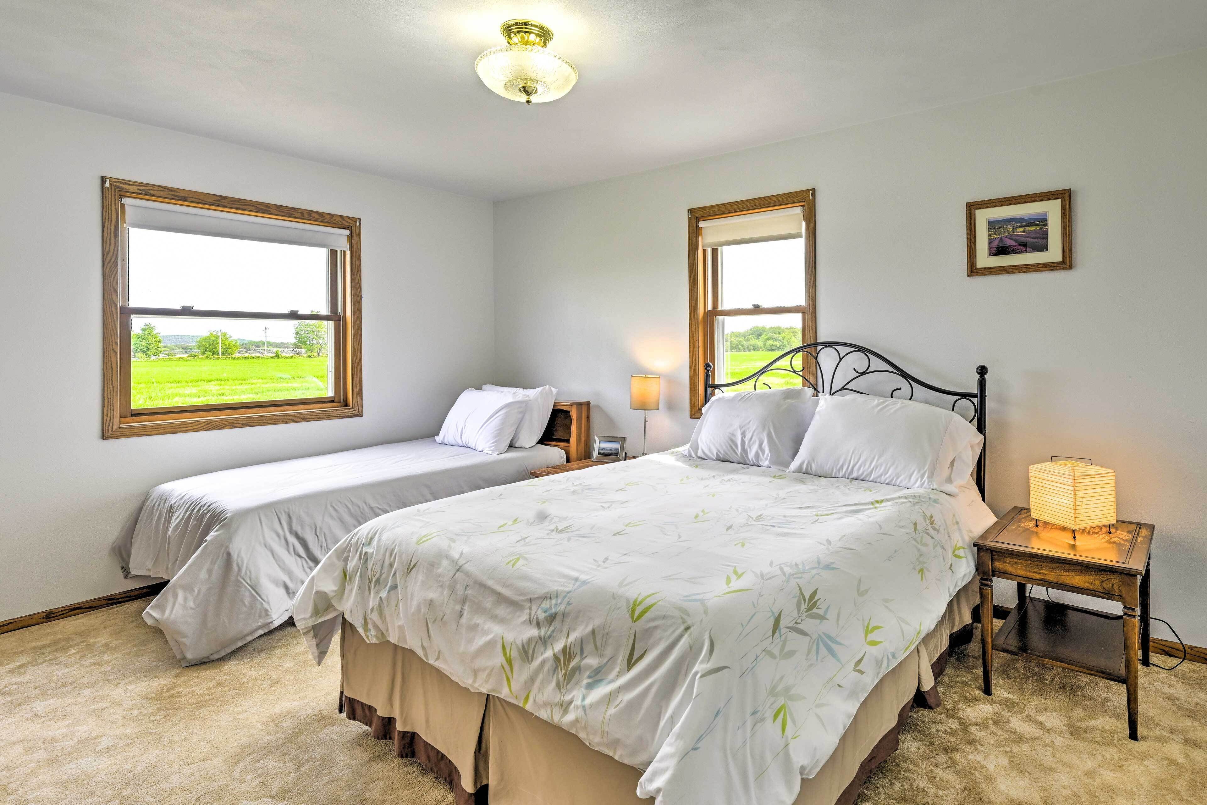 A bedroom with two beds, large windows, a bedside table with a lamp, and a framed picture on the wall.