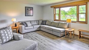A modern living room with a grey sectional sofa, two side tables with lamps, a wooden bench, and a large window with a view of greenery. - Stunning Wisconsin Retreat on Active Farm! (Arena)