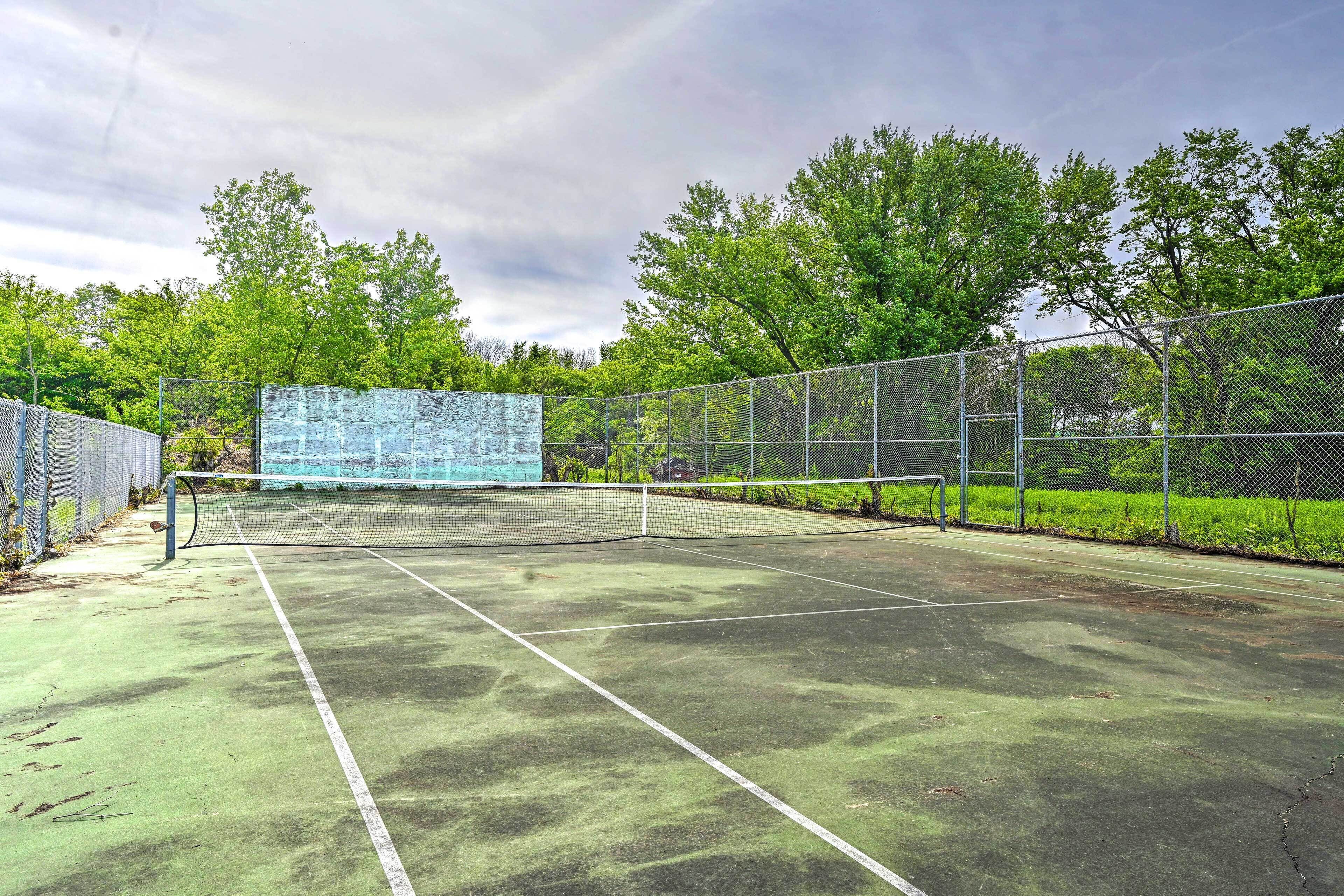 Una cancha de tenis al aire libre con red, rodeada por una cerca de malla y árboles.