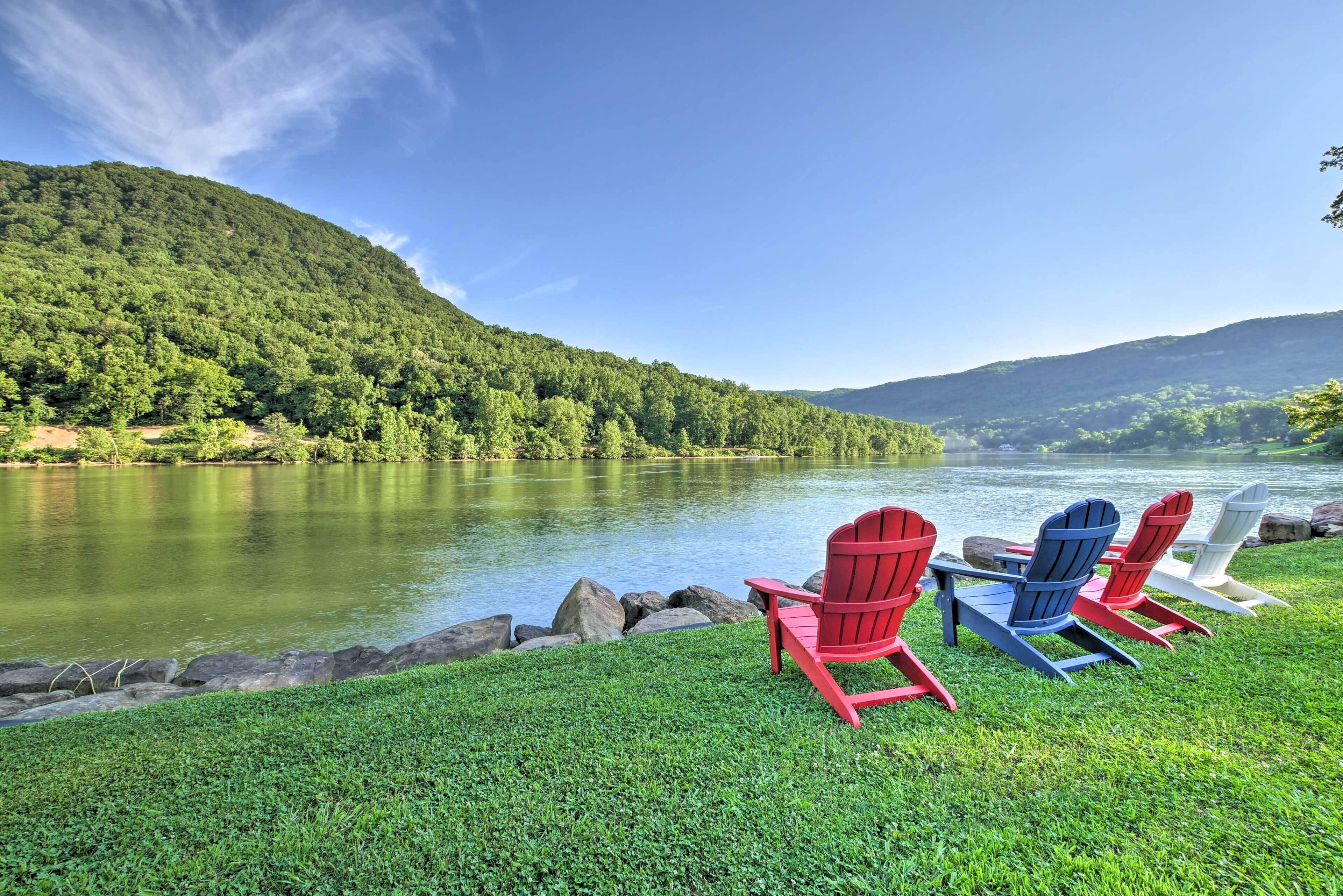 A lakeside view with a grassy area and three Adirondack chairs facing the water.