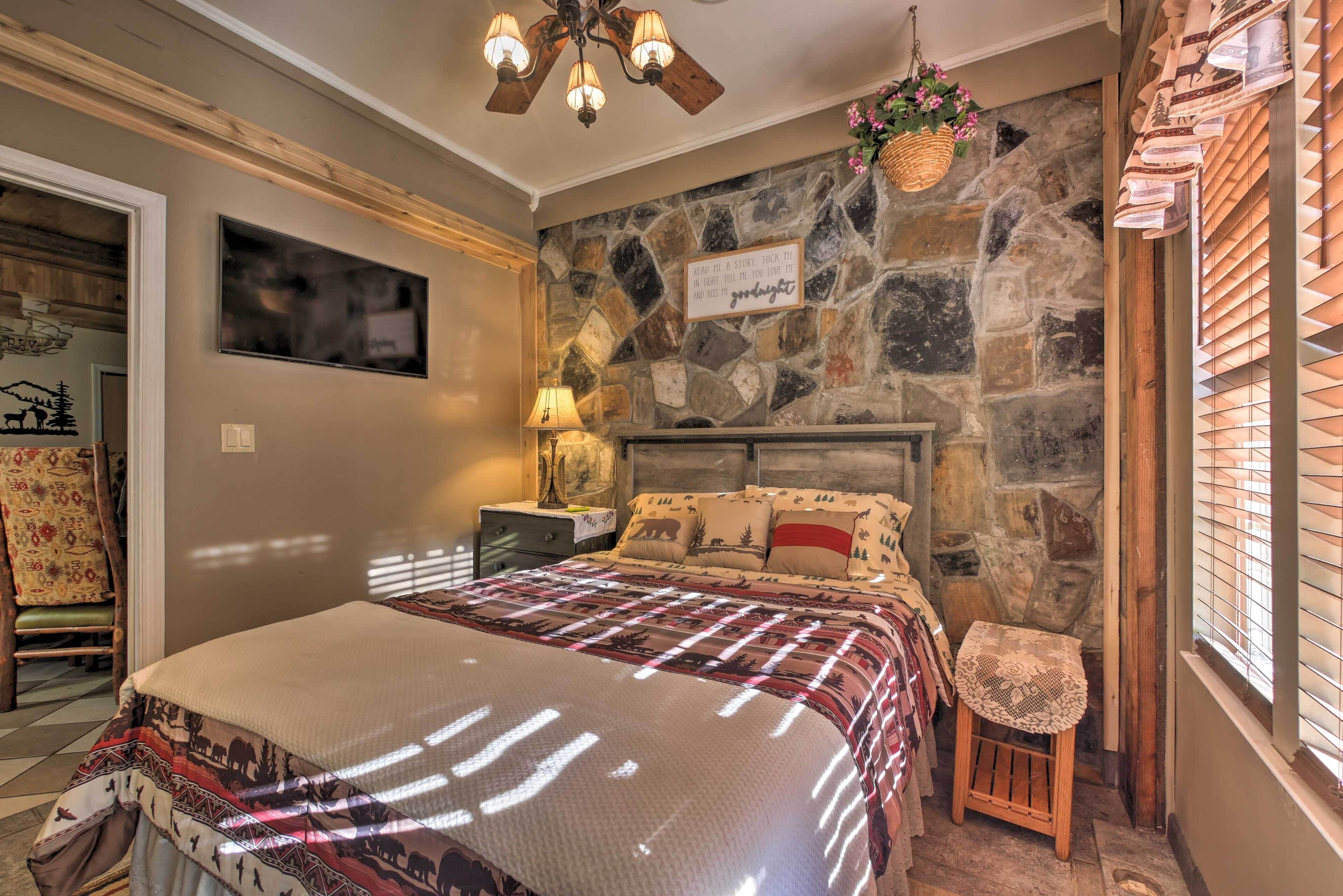 A cozy bedroom with a stone wall, a bed with patterned bedding, a nightstand with a lamp, and a small stool.