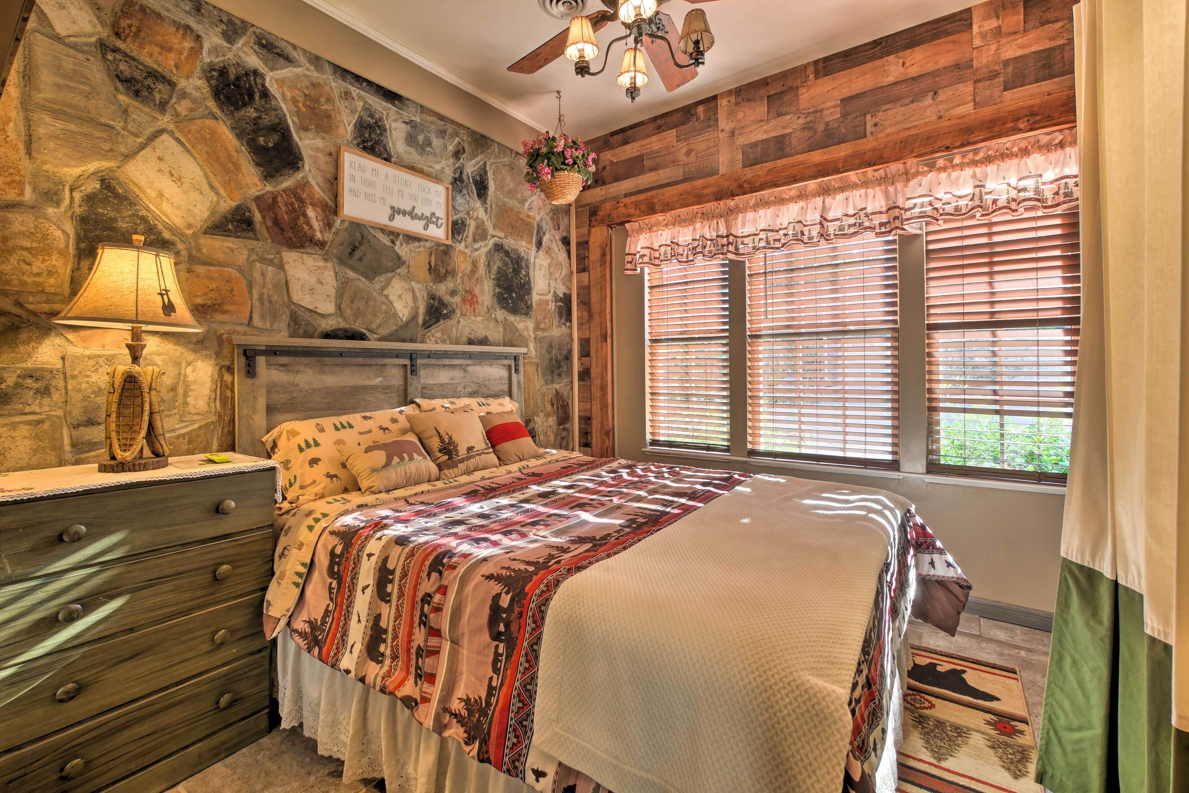 A bedroom with a stone wall, a bed with patterned bedding, a wooden dresser, and a window with blinds.