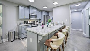 A modern kitchen with grey cabinets, a central island with bar stools, stainless steel appliances, and recessed ceiling lights.
