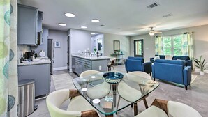 A modern living room with a glass dining table, blue sofas, and a kitchen area with grey cabinets and a central island.