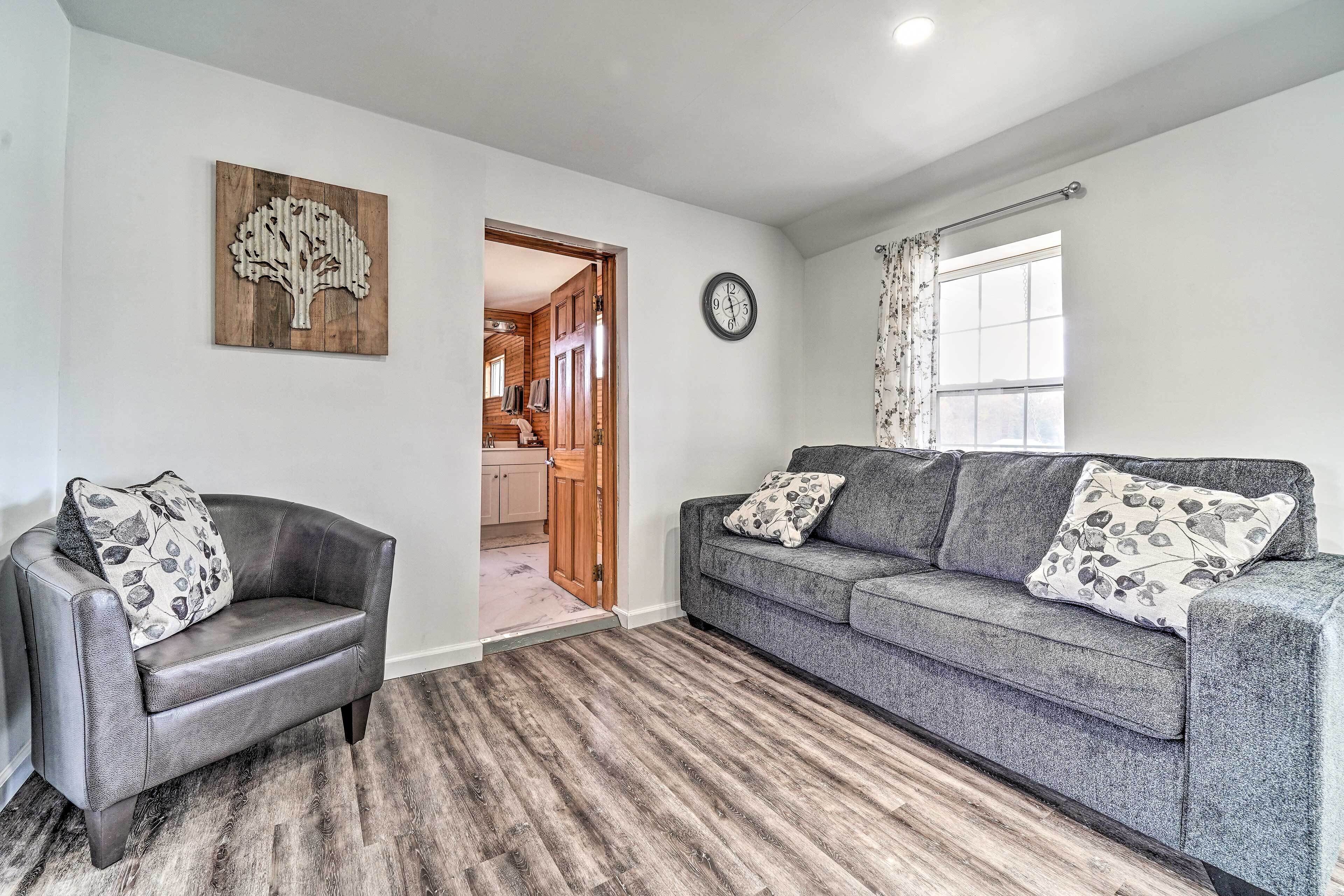 A living room with a grey sofa, a grey armchair, a wooden wall art piece, a clock, and a window with curtains.