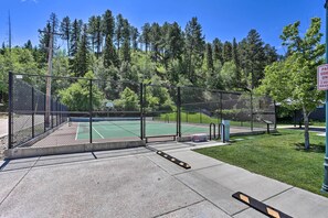 Una cancha de tenis cercada con superficie verde, rodeada de árboles y un área con césped.