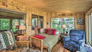 A cozy cabin interior with a wooden ceiling, a bed with red and patterned pillows, a blue armchair, and a window with a view of trees. - Year-round Waterfront Getaway: Lake Access + Dock! (South Haven)