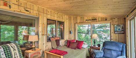 Interior acogedor de una cabaña con techo de madera, cama con almohadas rojas y estampadas, sillón azul y ventana con vistas a árboles.