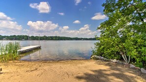 A sandy beach with a dock extending into calm water, surrounded by greenery. - Year-round Waterfront Getaway: Lake Access + Dock! (South Haven)