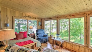 A cozy cabin interior with a bed, a chair, a table, and large windows offering a view of the outdoors. - Year-round Waterfront Getaway: Lake Access + Dock! (South Haven)