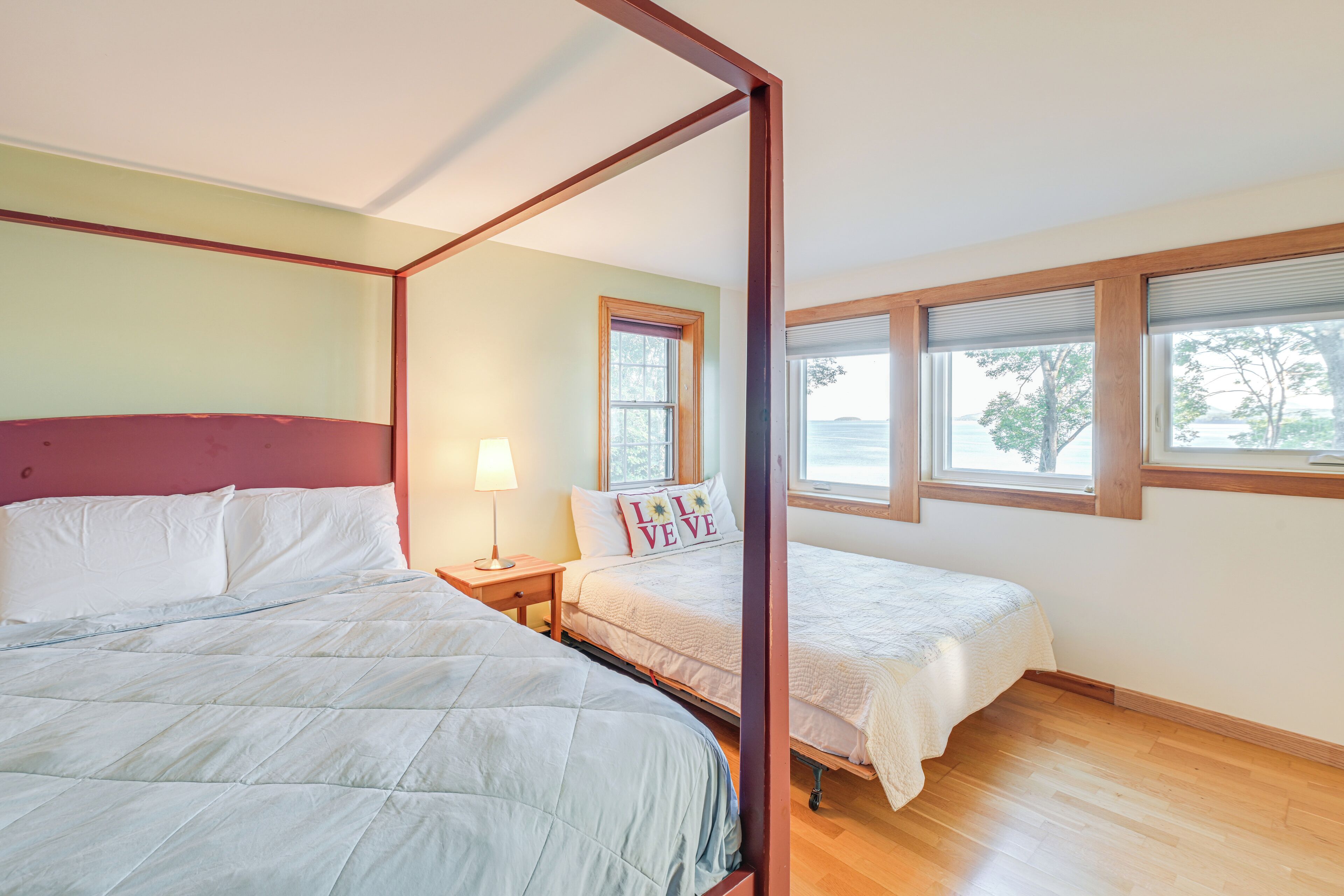 A four-poster bed with white bedding, a bedside table with a lamp, and a window with blinds.