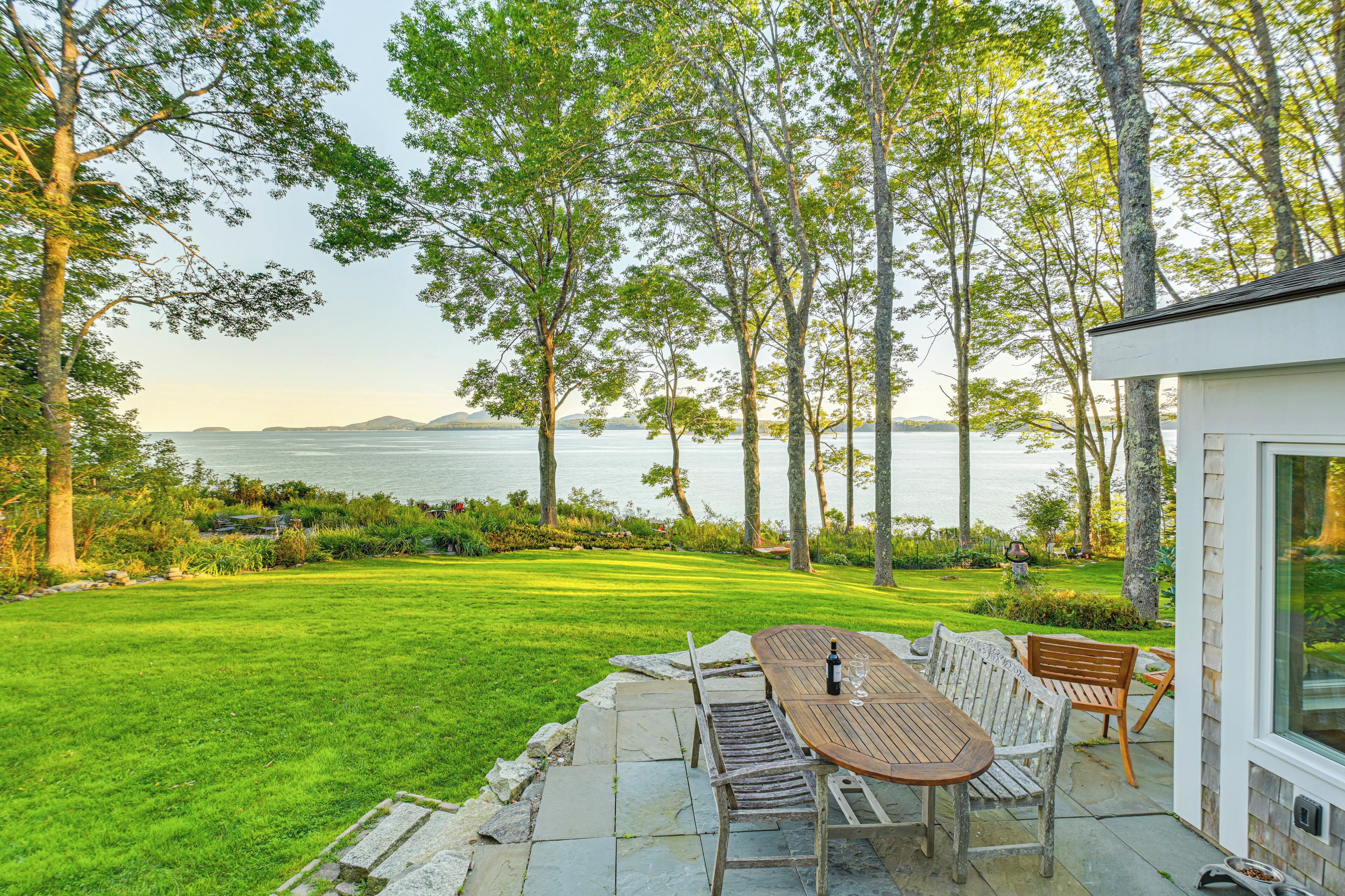 A patio with a table and chairs overlooking a body of water, surrounded by trees and a well-maintained lawn.