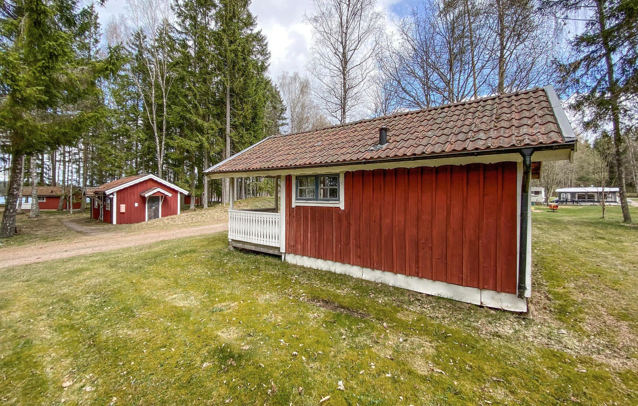 A red wooden cabin with a porch and a small window, surrounded by trees and a grassy area.