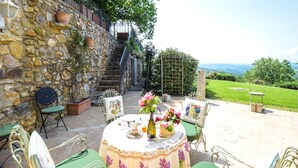 Eine Steinsäule mit einer Treppe, Sitzgelegenheiten im Freien mit grünen Stühlen und einem Tisch, der mit Blumen gedeckt ist, und ein Blick auf eine grüne Landschaft.