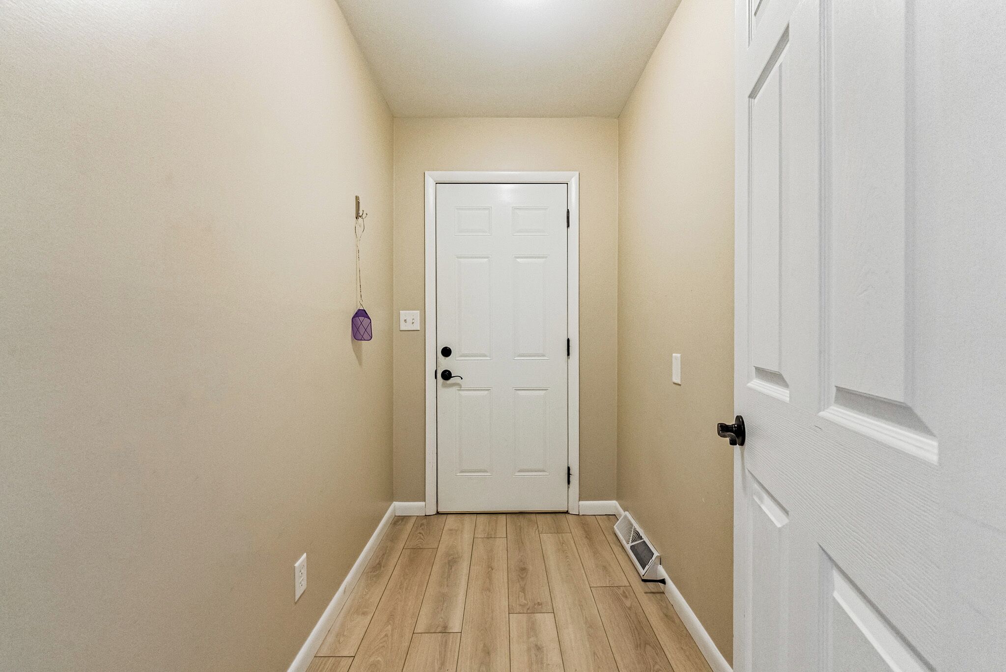 Un couloir étroit, avec une porte blanche au fond, des murs de couleur beige et un plancher en bois.