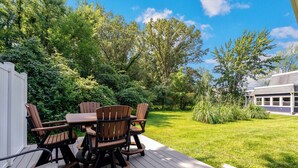 A deck with a table and chairs, surrounded by lush greenery and a house in the background. - Winona Lake Access With Pier (Warsaw)
