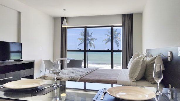 Ein Hotelzimmer mit Bett, Fernseher, Esstisch für zwei Personen und Meerblick durch große Fenster.
