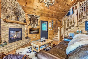 A log cabin interior with a stone fireplace, a mounted fur head, and a wooden staircase.