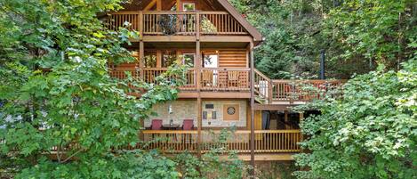 A multi-story log cabin with a balcony, surrounded by dense forest.