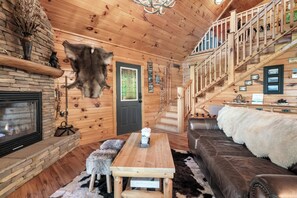 A log cabin interior with a stone fireplace, wooden walls, a leather sofa, a wooden table, and a staircase.