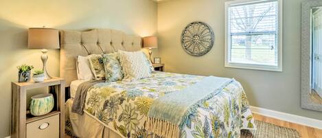 A bedroom with a large bed, two bedside tables, a ceiling fan, a window with blinds, and a decorative wall piece.