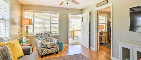 Un salon avec un canapé gris, un fauteuil à motifs et un ventilateur de plafond. Une pancarte indique : « MAISON AU BORD DU LAC ».
