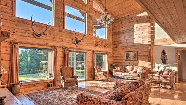 A spacious log cabin interior with large windows, a chandelier, and rustic decor including antlers. - Brookings Vacation Rental Lodge on 88 Acres! (Brookings)