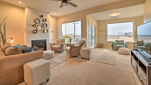 A spacious living room with a fireplace, comfortable seating, large windows, and a ceiling fan. - Chic Manistee Cottage: Steps to Lake Michigan! (Manistee)
