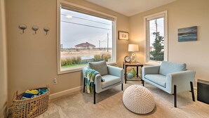 A living room with two light blue armchairs, a round ottoman, a wicker basket, and a large window with a view of a building and greenery. - Chic Manistee Cottage: Steps to Lake Michigan! (Manistee)