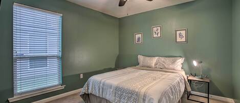 Un dormitorio con cama, ventilador de techo, mesita de noche con lámpara y ventana con persianas.