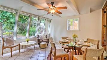 A dining area with a glass table, wooden chairs, a ceiling fan, and large windows offering a view of greenery.
