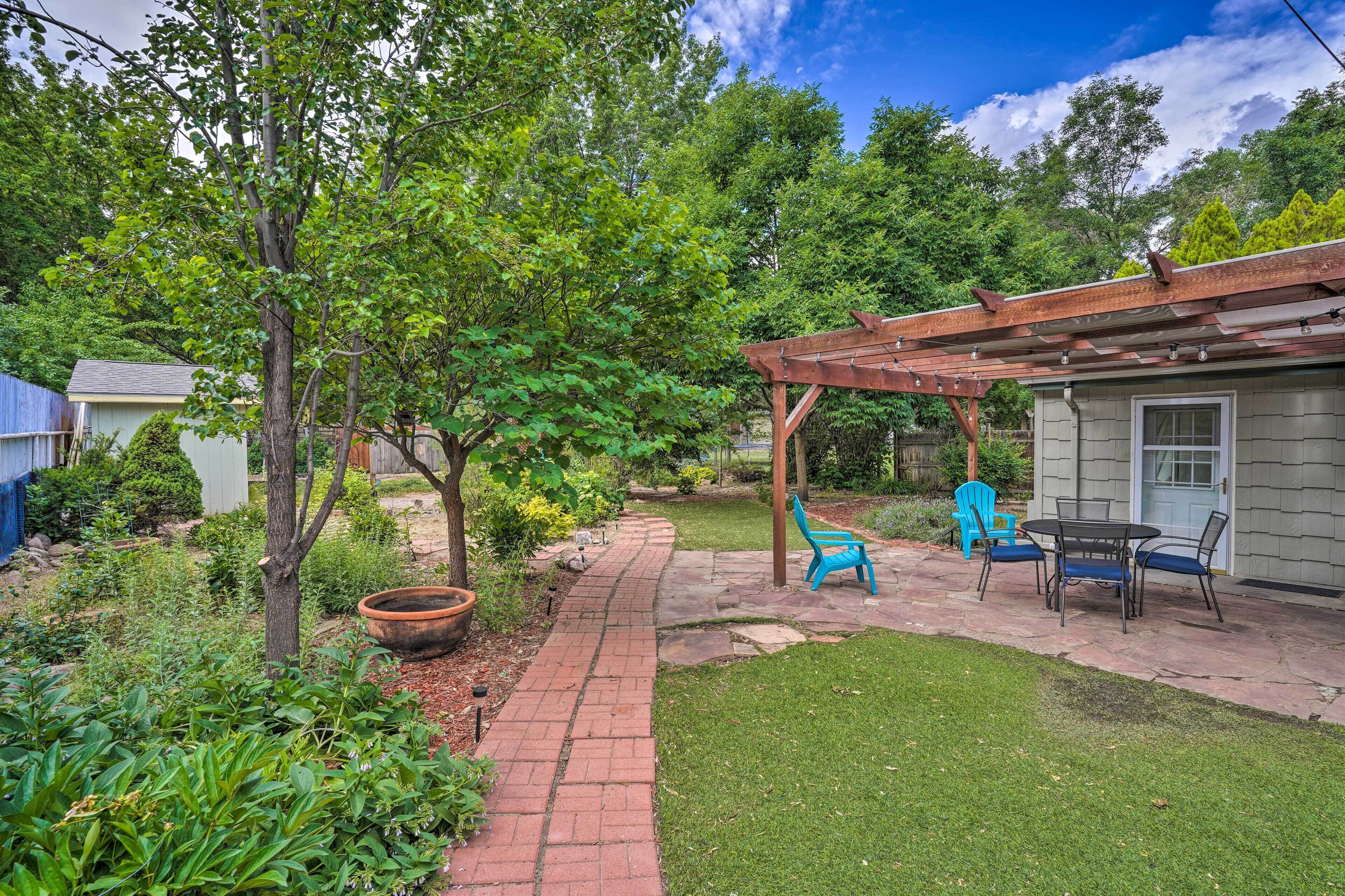 A backyard with a brick pathway, a wooden pergola, a table with chairs, and a blue chair.