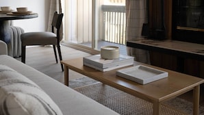 Ein modernes Wohnzimmer mit einer Couch, einem Couchtisch mit Buch und Kerze, einem Sessel und Blick auf einen Balkon mit Geländer.