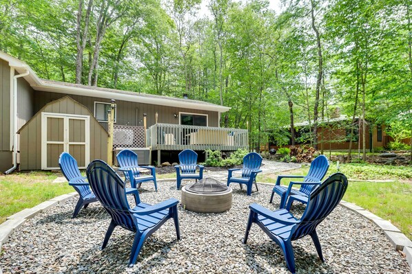 Eine Terrasse mit Adirondack-Stühlen und einem Feuerkorb, umgeben von Kiesflächen und Bäumen.