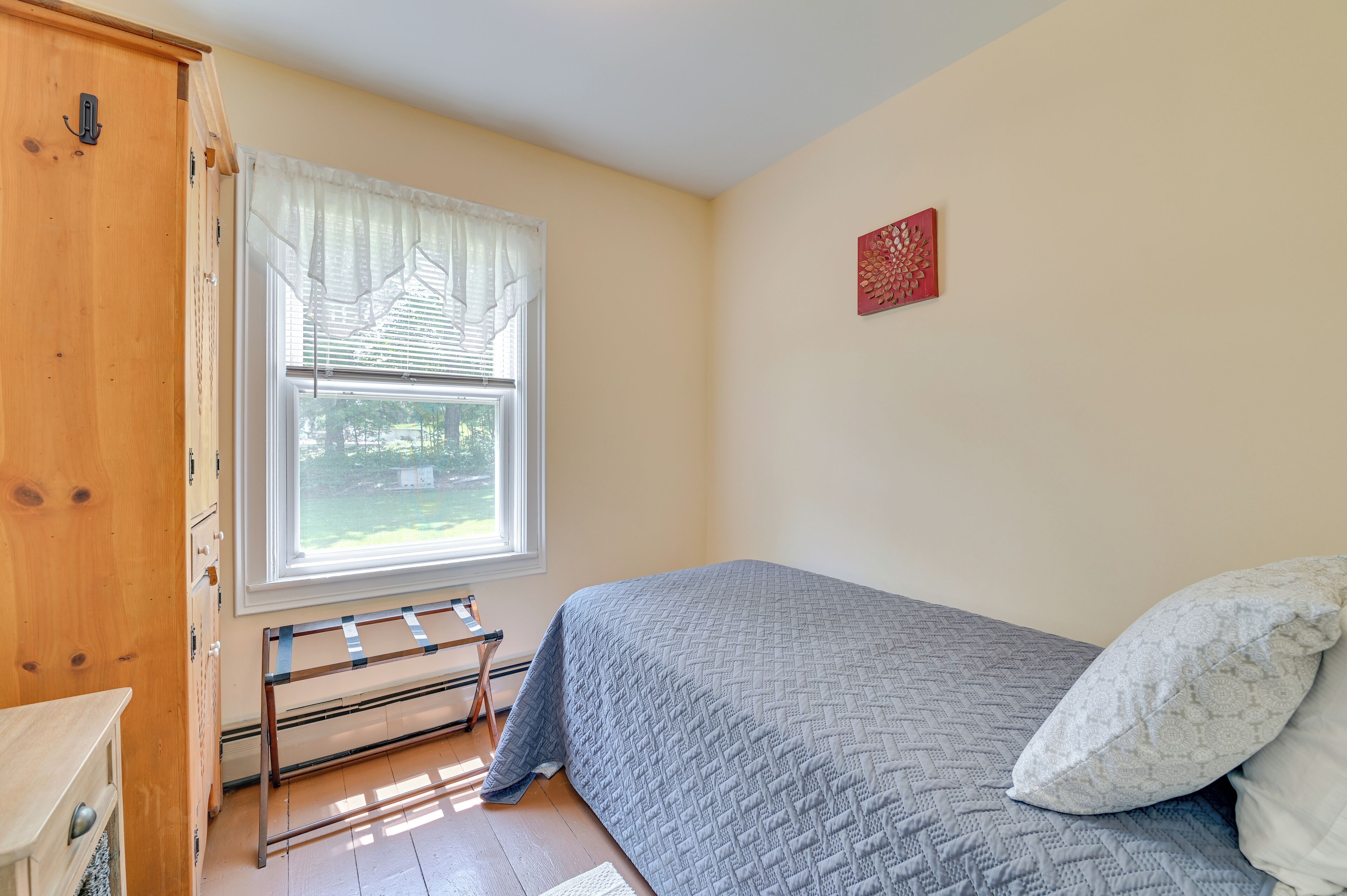 A bedroom with a bed, a window with curtains, a wooden wardrobe, and a wall-mounted artwork.