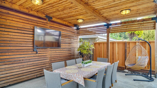 Une terrasse extérieure dotée d’un plafond en bois, d’une table recouverte d’une nappe à motifs, de chaises en osier, d’un fauteuil suspendu et d’un téléviseur fixé au mur.