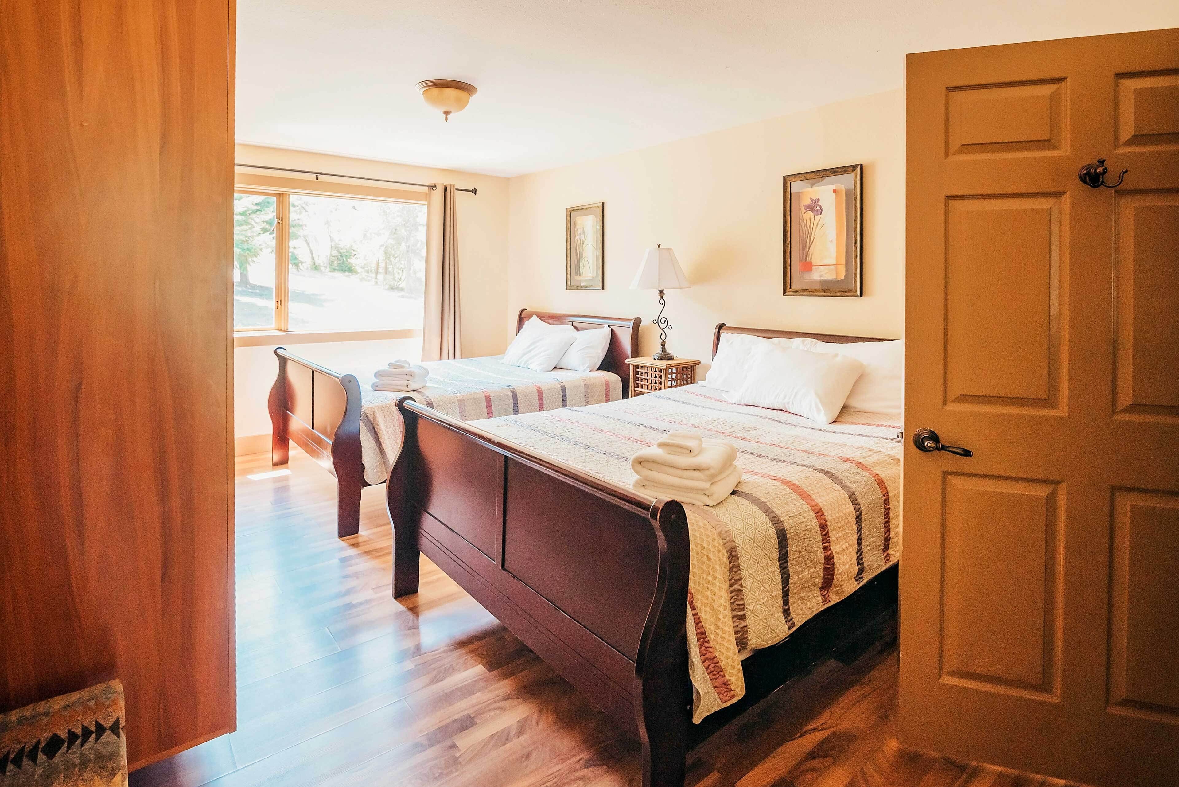 A hotel room with two wooden beds, a window with curtains, a door, and a framed picture on the wall.