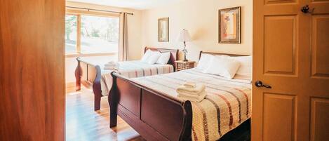 A hotel room with two wooden beds, a window with curtains, a door, and a framed picture on the wall.