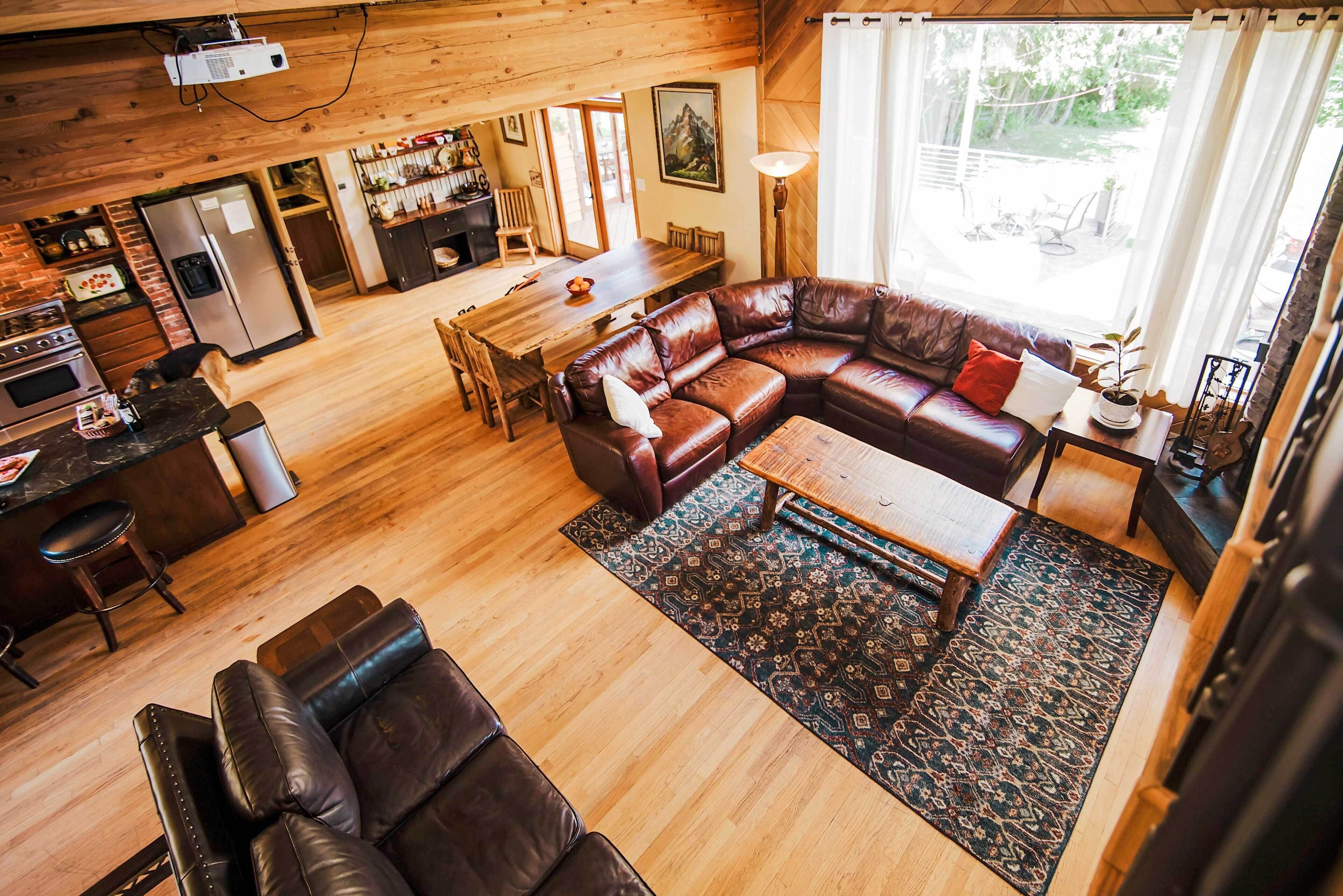 L’intérieur chaleureux d’un chalet avec une table à manger en bois, des canapés en cuir, un coin cuisine et une grande fenêtre agrémentée de rideaux.