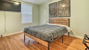 A bedroom with a bed, wooden floor, a window with blinds, and two framed pictures on the wall.