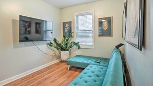 A living room with a teal sofa, a flat-screen TV mounted on the wall, a potted plant, and framed artwork.