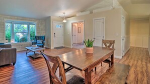 A spacious living room with a wooden dining table, two chairs, a sofa, and a coffee table. There are large windows with a view of greenery outside. - Beautiful Pet-friendly Washougal Home w/ Fire Pit! (Washougal)