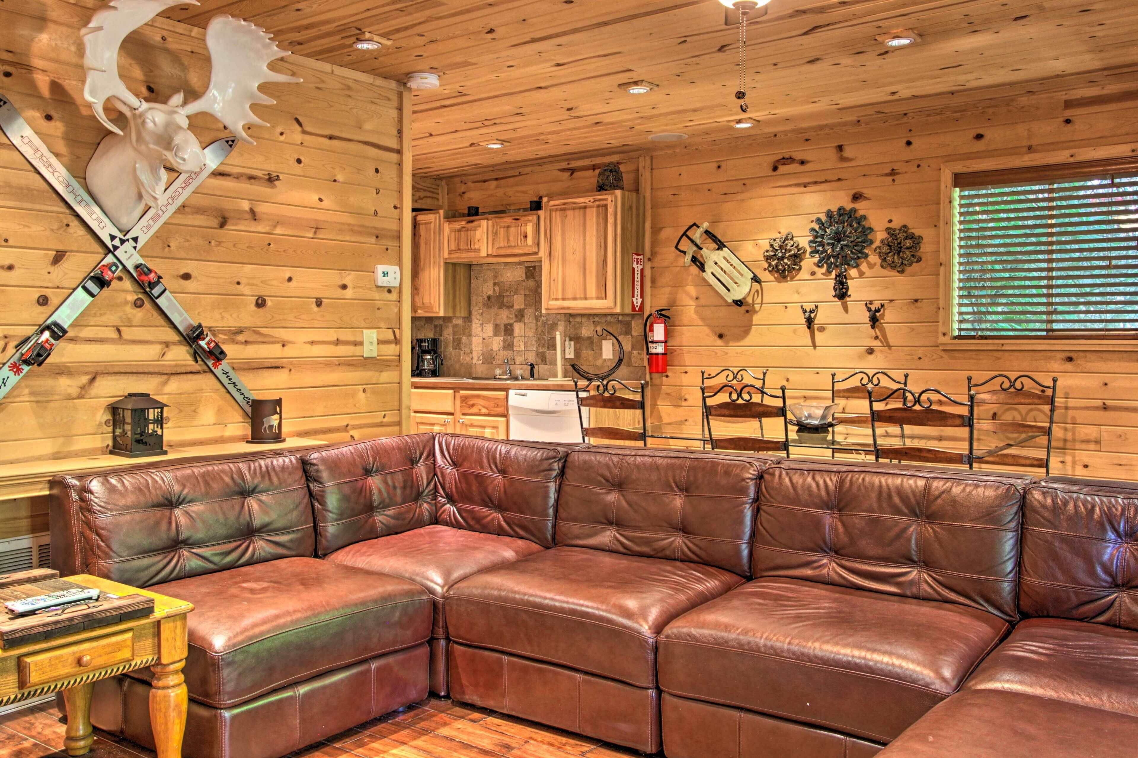 A cozy cabin interior with a leather sectional sofa, wooden walls, a kitchen area, and a mounted axe.