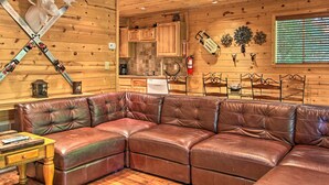 A cozy cabin interior with a leather sectional sofa, wooden walls, a kitchen area, and a mounted axe. - Rustic Gatlinburg Chalet w/ Hot Tub: 2 Mi to Town (Gatlinburg)